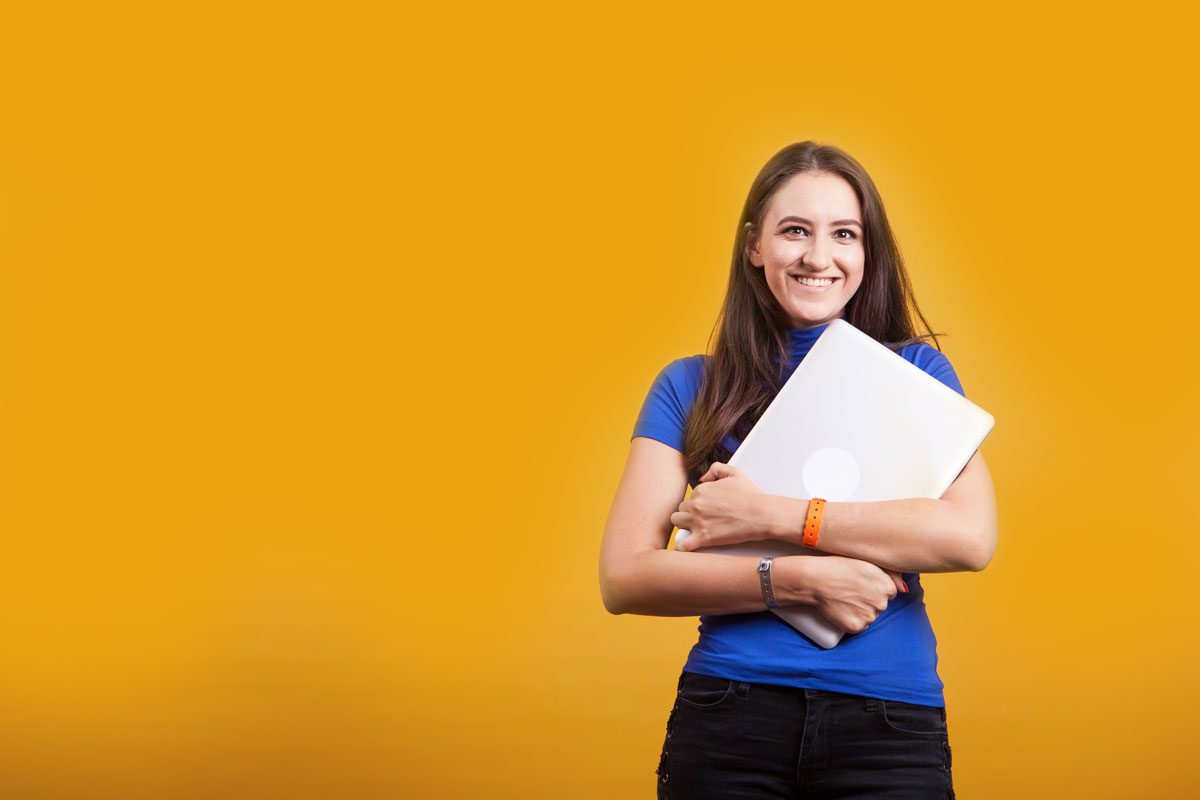 A smiling young woman in a blue shirt hugging a laptop against a gold background.