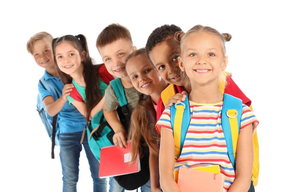 A row of smiling elementary students with backpacks leaning into the frame.