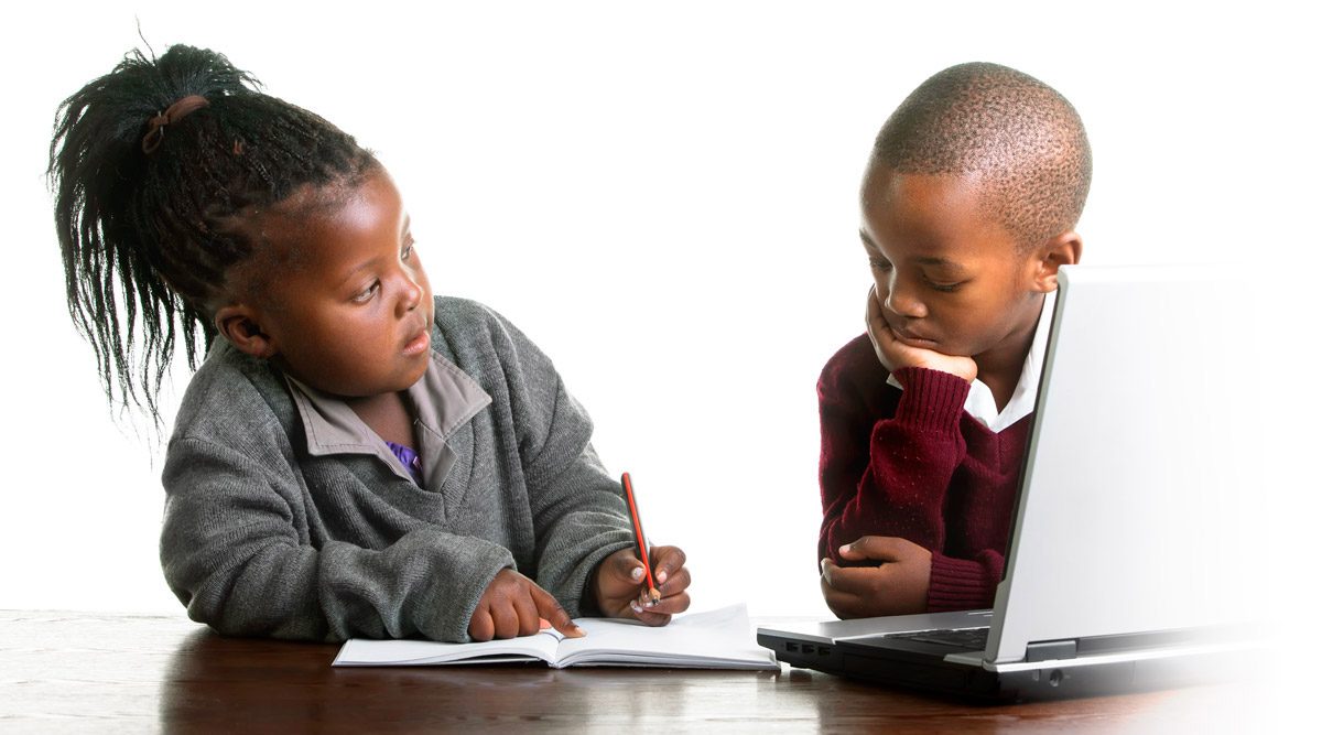 Two young students working at a table, one writing in a notebook and one using a laptop
