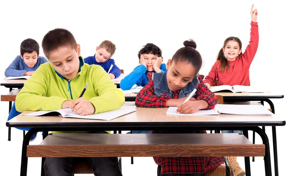 Elementary students writing in notebooks at classroom desks while a girl raises her hand