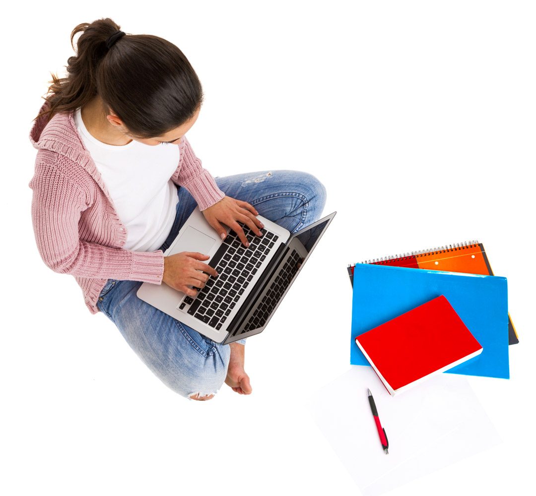 A girl sitting cross-legged typing on a laptop beside a stack of colorful notebooks.