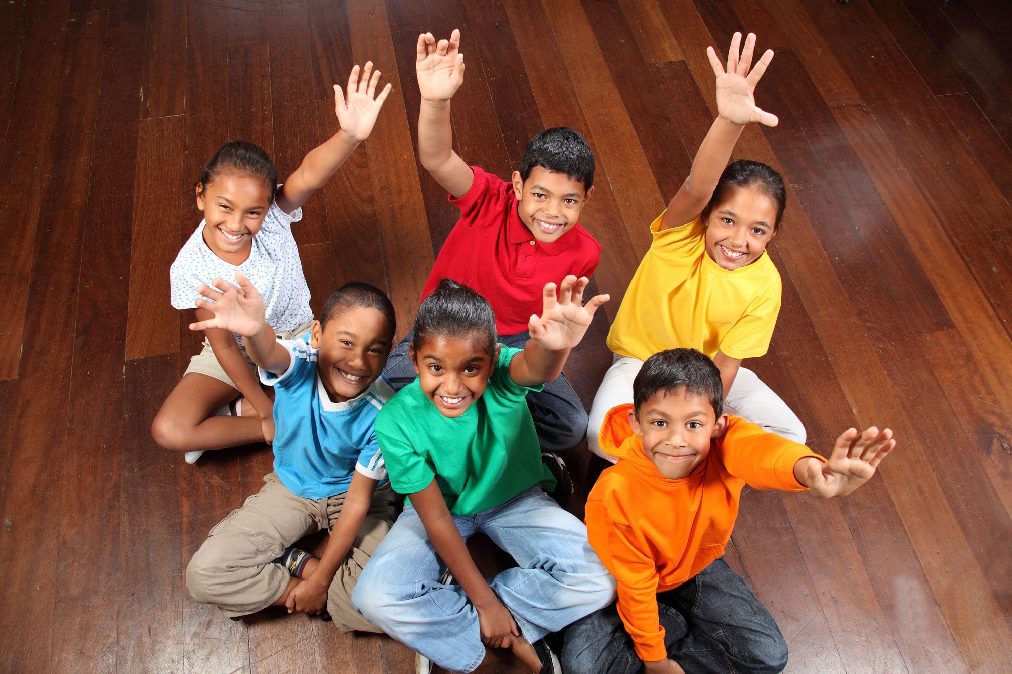 A group of smiling elementary students sitting on a wooden floor, waving their hands in the air.